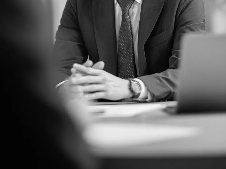 グレーのスーツとネクタイを着用した人物が、デスクで両手を組み相談に乗っている