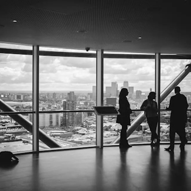 高層ビルの大きな窓際で、都会の街並みを背景に3人のビジネスパーソンがシルエットで立ち話をしている様子